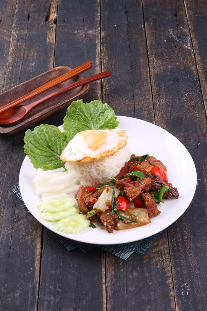 brand-01 Thai basil chicken with rice, fried egg, and fresh vegetables on a rustic wooden table.