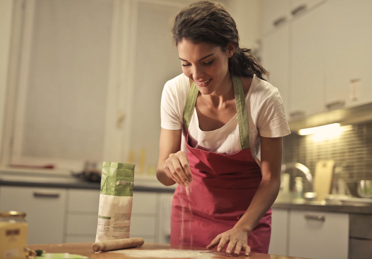 digital-01 Low angle of positive young female in apron sprinkling flour over table while preparing food in modern light kitchen