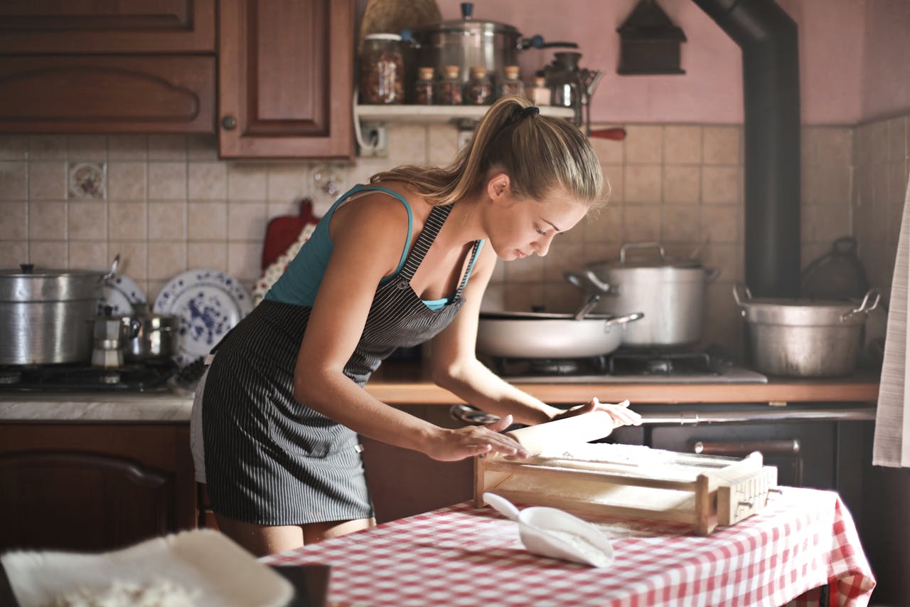 Services-02 Side view of housewife wearing apron standing at table in cozy kitchen and preparing dough for baking while using rolling pin