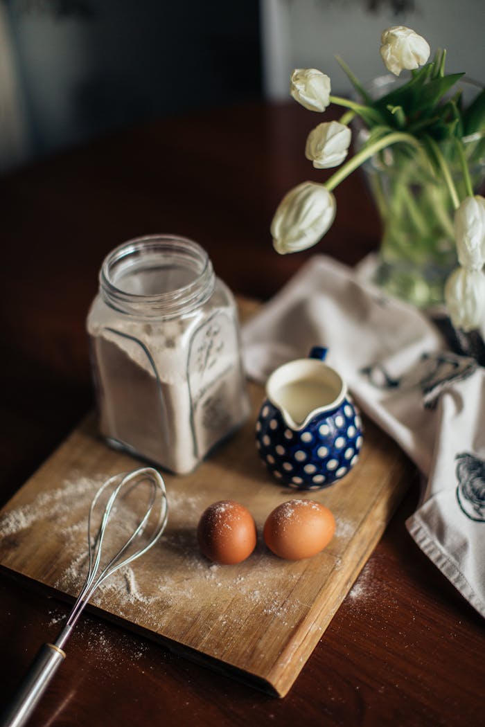 creative-03 From above of vase with white flowers placed near fresh milk eggs and glass jar with flour on cutting woody board at kitchen