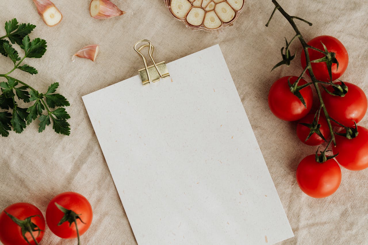 Services-03 From above of blank clipboard with golden paper binder placed on linen tablecloth among tasty red tomatoes on branches together with cutted garlic and green parsley devoted for recipe or menu placement