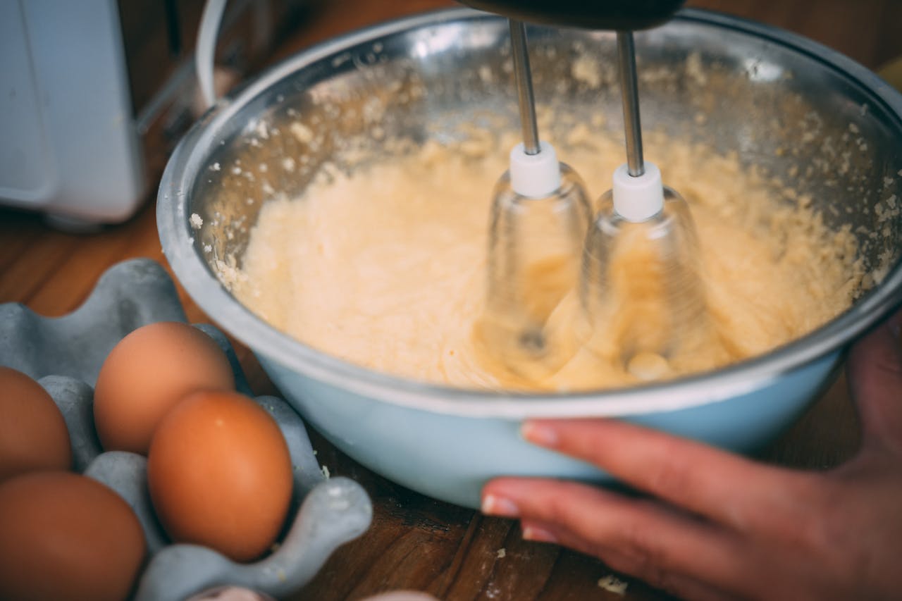 digital Close-up of batter being mixed in a bowl with eggs nearby on a wooden table.