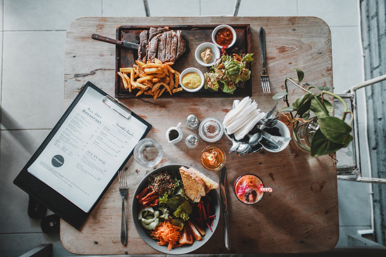 why-choose-us Top view of wooden table with salad bowl and fresh drink arranged with tray of appetizing steak and french fries near menu in cozy cafe